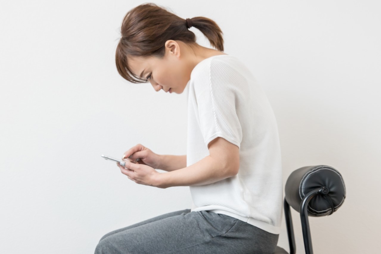 猫背でスマホをいじっている女性のイメージ。肩こりや筋緊張による痛みを表現した画像